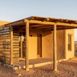 Maison désertique bioclimatique aux murs enduits intégrant des bouteilles remplies de sable et une pergola d’ombrage, éclairée par une lumière chaude.