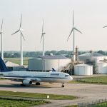 Avion de ligne stationné devant une unité industrielle de carburant durable avec réservoirs, panneaux solaires et éoliennes.